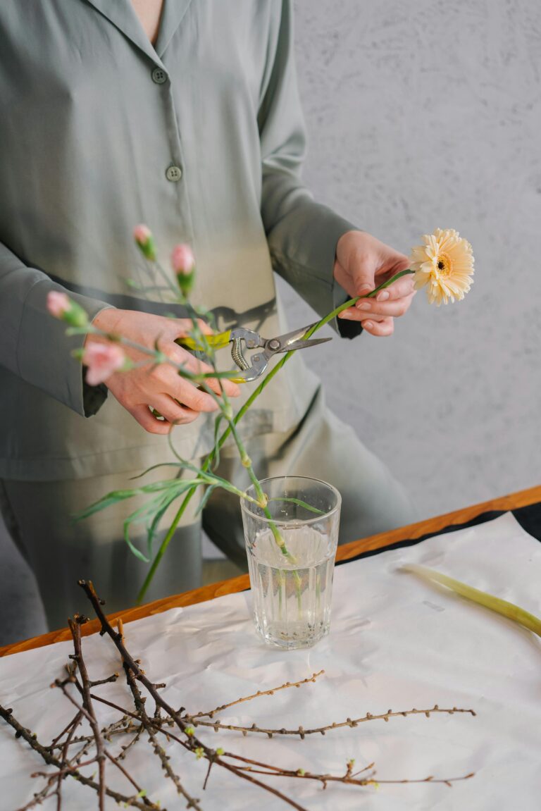 A woman in green outfit trimming flowers for a floral arrangement indoors.